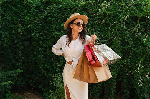 Happy young woman wear white clothes and straw hat hold package bags after shopping isolated on greenery background. Summer rest concept. Copy space