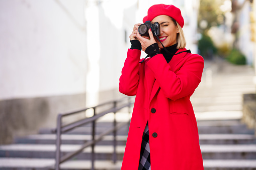 Attractive female photographer in stylish clothes taking photo on modern photo camera while standing on stairs near building on blurred background