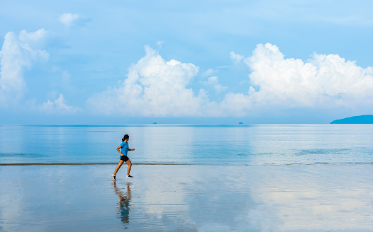 Woman running along the beach of the sea with beautiful sky and blue sea background, healthy lifestyle outdoor exercise concept.