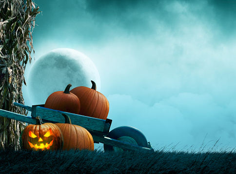 A jack o'lantern rests in front of a wheelbarrow full of pumpkins in front of a full moon rising over a grassy hill.