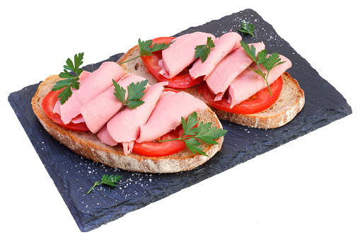 Two sandwiches on black slate board. Cooked meal isolated on white background. Mortadella sliced on tomatoes with rye bread and parsley.