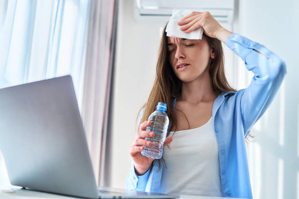 schwitzende frau, die unter warmem wetter und durst leidet, wischt sich die stirn mit einer serviette ab und kühlt sich mit einer kalten wasserflasche ab, während sie an einem heißen sommertag online am computer arbeitet - air conditioner cold home interior climate stock-fotos und bilder