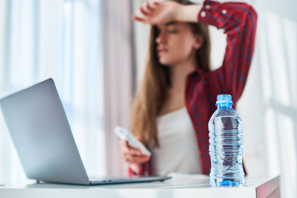 verärgerte müde freiberuflerin, die unter hitze und heißem wetter leidet, kühlt sich mit kalter erfrischungswasserflasche ab, während sie am sommertag online am computer arbeitet - air conditioner cold home interior climate stock-fotos und bilder