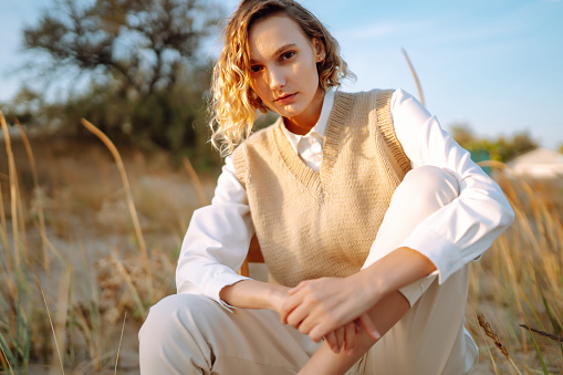 Slender model with windy hair in stylish clothes poses among the wild grass on the beach. Beautiful woman standing on the windy coast. Freedom. Concept of fashion, relaxation, recreation.