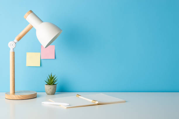 istock Get ready to learn! Side view photo of a desk adorned with school supplies, open notebook, flowerpot, lamp against a blue wall with sticky note paper. Ample space for text or ads 1595793476