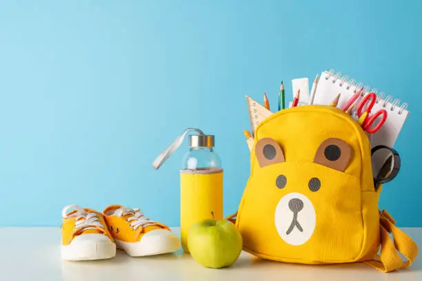 Photo of Classroom theme. Side view photo of desk adorned with school essentials - pencils, ruler, album for drawing, and more. Charming teddy-bear schoolbag, shoes and apple, water bottle add delightful touch