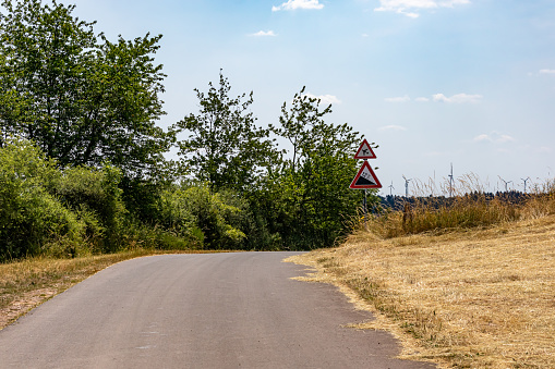 Country road between dry grass and wild vegetation, traffic signs: airport warning or low flying airplane and steep 12 percent downhill gradient in background, sunny summer day in Utscheid, Germany