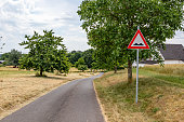 Country road with a traffic sign indicating speed bump, between dry grass, farmland and farms