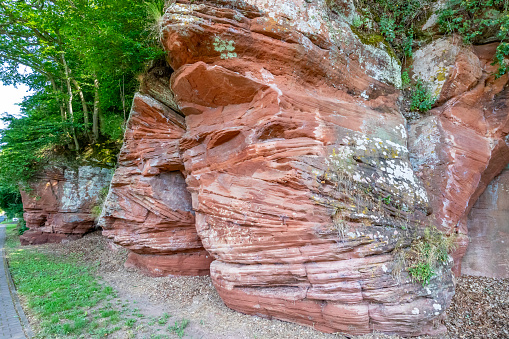 Red sandstone rock formation, irregular texture, mold, white spots and linear erosions caused by passage of time, trees with green foliage in background, sunny day around Stausee Bitburg lake, Germany