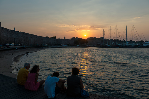 People watching the sunset on the beach with a view of the harbor and the city walls. August 5, 2016 Old Town, Rhodes Island, Greece