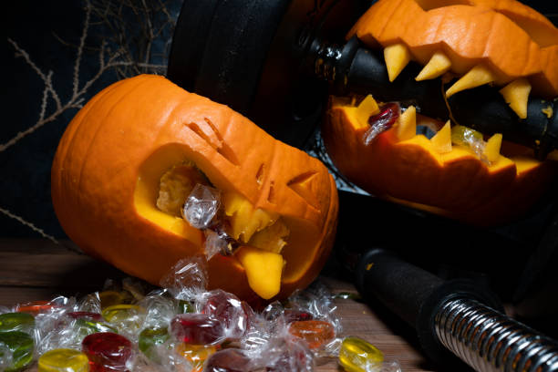 istock Halloween pumpkin clenching teeth on barbell dumbbell, crushing other carved Jack-o'-lantern. Gym workout sport training concept. Healthy diet autumn fall composition. 1595925607