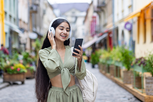 Young beautiful id woman walks in the city, tourist holds phone in hands, uses headphones to listen to music and online radio podcasts, dances and sings along happily.