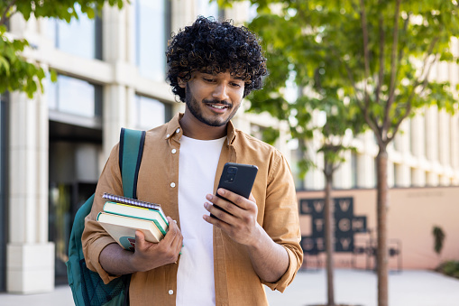Young Indian male student using mobile phone, standing outside on street, holding books in hand, reading message, looking for campus, waiting for meeting.