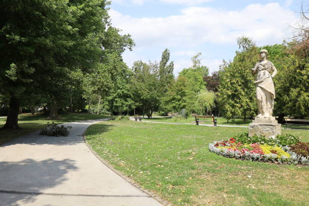Tranquil park-like setting filled with an assortment of trees and a lush grassy area. Prominent large trees with vibrant green foliage are scattered throughout the scene, while one tree on the left side of the image hints at the arrival of autumn with its leaves beginning to turn brown. This image was taken in Ambronay village, in the town hall public park, in Ain department, Auvergne-Rhone-Alpes region in France.