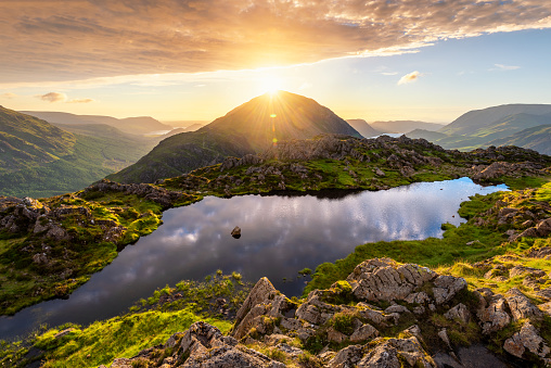 Beautiful majestic landscape with sun shining through mountain peak at Haystacks Tarn in the Lake District, UK. Breathtaking scenic viewpoint with calm reflections in body of water.
