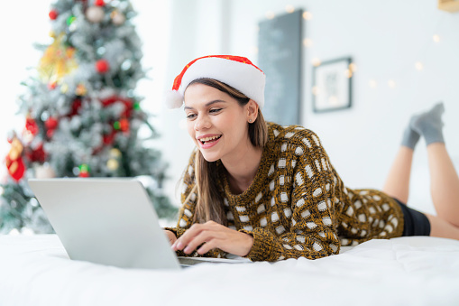 Beautiful woman lies comfortably on her bed, surrounded by the enchanting ambiance of Christmas decorations. Wearing cheerful Santa hat and exudes happiness while engaging in modern holiday activities using her laptop and wireless technology. Combining the pleasure of relaxation and enjoyment with long holiday.
