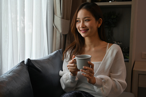 Happily of Young Asian woman focus out of the window and holding a cup of coffee while sitting on the couch.