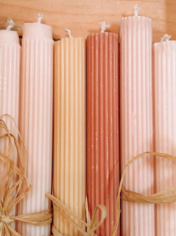 Vertical high angle closeup photo of a group of long pink, gold and orange coloured candles with a rib pattern, wrapped two together with raffia straw, on display on a wooden bench in a gift shop.