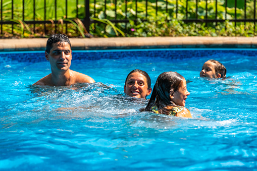 A couple with their two daughters having fun and swimming in their back yard pool during a sunny day of summer.