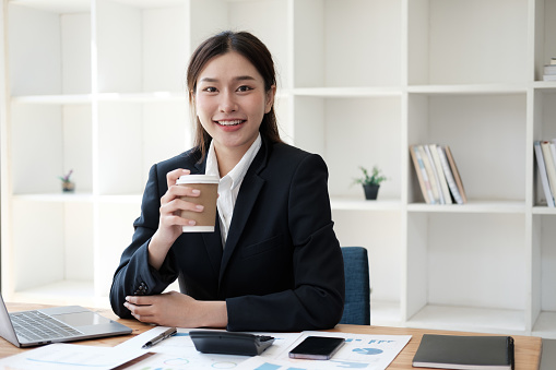 Young adorable Asian businesswoman holding a coffee cup while sitting at her office desk. taking a break from working.