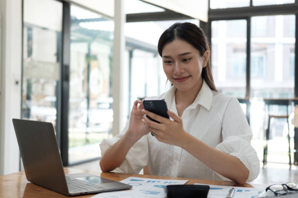 istock Young beautiful Asian businesswoman using smartphone, cheating with her friends while working in the office room. 1596266852