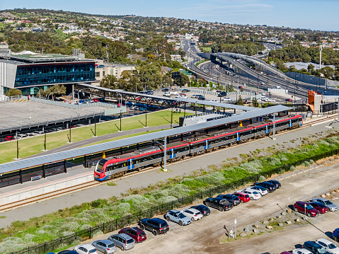 Essential & efficient rail infrastructure: aerial view modern electric train entering university terminus station after exiting bridge over wide, multi-lane highway with elevated interchange and overpasses in treelined suburb. Train line links university campus, public and private hospitals with city and suburbs. Flinders Rail Link with covered walkway to Flinders Hospitals, Southern Expressway, Main South Road  all visible with Darlington service centres in background. Logos & ID edited.