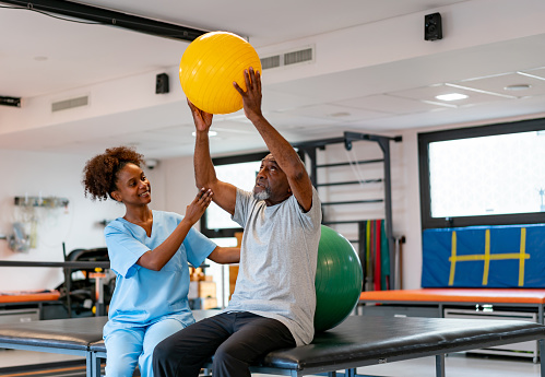 Cheerful physiotherapist helping senior black patient workout with a fitness ball at a rehabilitation center  - healthcare concepts