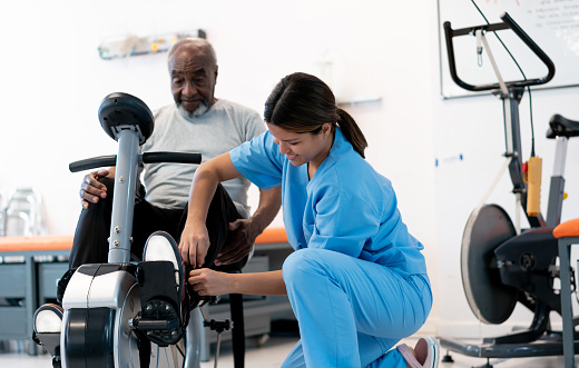 Female physical therapist adjusting the straps on her senior's patients feet before starting to exercise on a static bicycle - Healthcare concepts