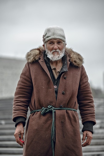 Portrait of an old homeless man with gray beard in brown coat and white hat. medium plan. looking away.