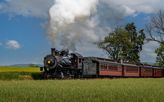 A vintage locomotive train emits a plume of thick, white smoke as it chugs along a set of railway tracks, passing by a lush green field in
