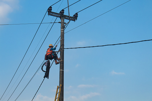 Banjar, Indonesia - August 3, 2023: Electricity concept, Electricians Technician with hardhat and safety uniform checking repairing fixing electric wire on power pole. High voltage power lines and blue sky background.