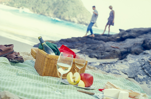 Close-up view of wicker picnic basket, sandwich, fruits and bottle of wine with glasses on napkin