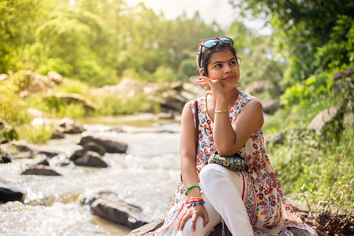 A pretty beautiful Indian woman sitting besides a rocky river in green background