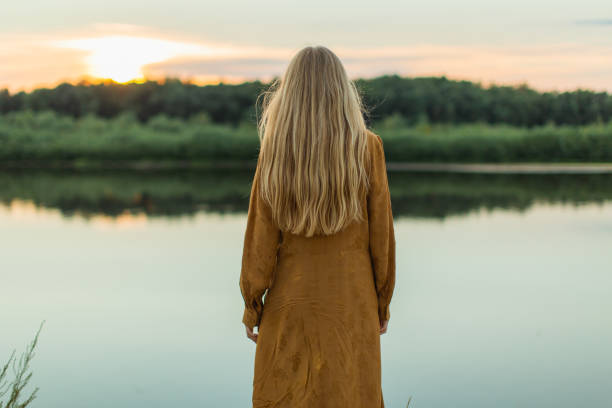 veduta posteriore. donna bionda in vestito giallo che riposa sulla riva del fiume. giorno d'estate. capelli lunghi. sabbia. - long long hair dress young adult foto e immagini stock