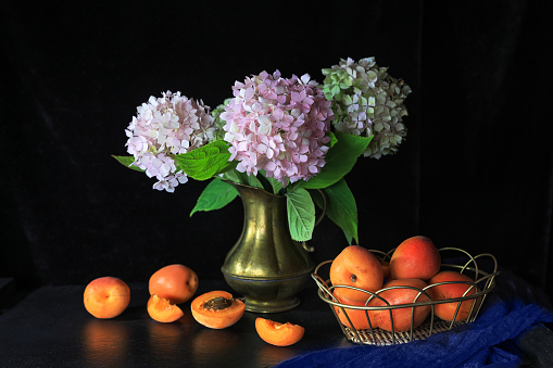 Fresh apricots in metal basket on table with flowers in bronze vases against dark background - dark and moody photography