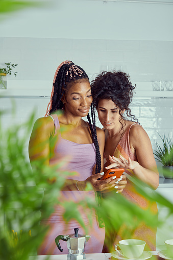 Two female friends scrolling smart phone, having fun in the modern white kitchen, using dating app, on line shopping, digital payment, messaging, reading news and gossip, looking at new products, representing a healthy lifestyle and friendship, wellbeing, bonding, joy and togetherness