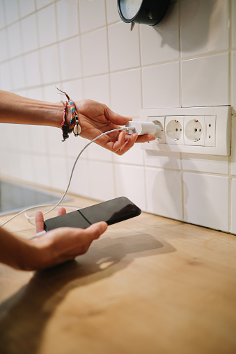 Woman using a wall charger with a cable in the kitchen to charge her smartphone.