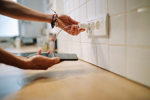 Woman using a wall charger with a cable in the kitchen to charge her smartphone.