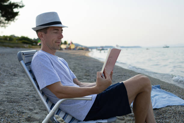istock Man with coffee cup is reading book while he is sitting in deck chair on the beach. 1596810734