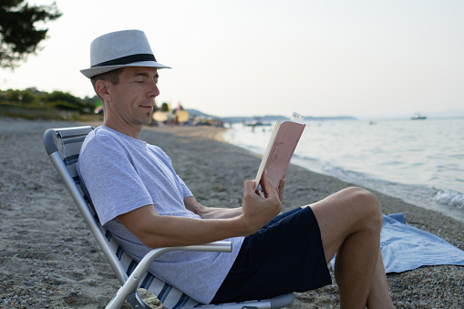 Man with coffee cup is reading book while he is sitting in deck chair on the beach.