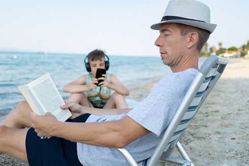 Boy with headphones is listening music while his father is reading book in deck chair on the beach.