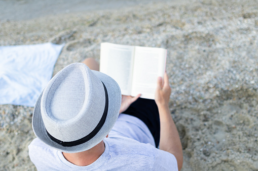 Man with a hat is reading book on the beach.
