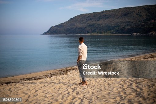 istock Young man in a relaxed pose on a sandy beach, with a tranquil ocean in the background 1596820801
