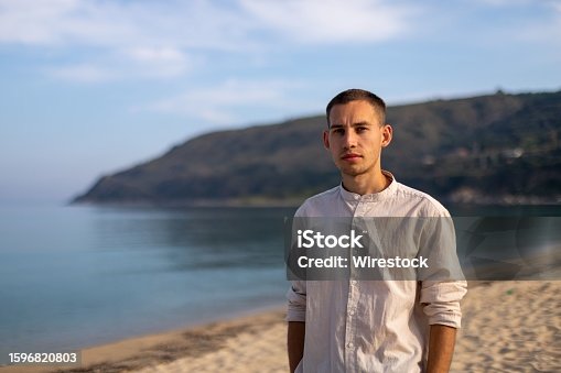 istock Young man in a relaxed pose on a sandy beach, with a tranquil ocean in the background 1596820803