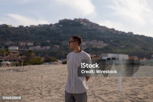 istock Young man in a relaxed pose on a sandy beach, with a tranquil ocean in the background 1596820808