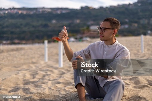 istock Young man in a relaxed pose on a sandy beach, with a tranquil ocean in the background 1596820813