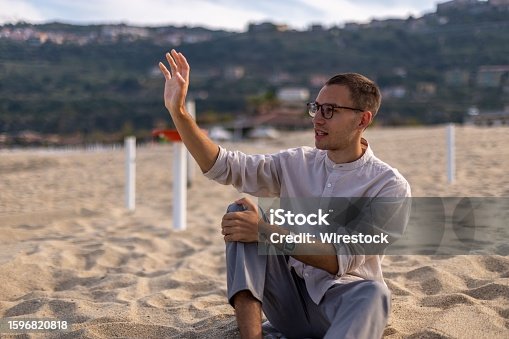 istock Young man in a relaxed pose on a sandy beach, with a tranquil ocean in the background 1596820818