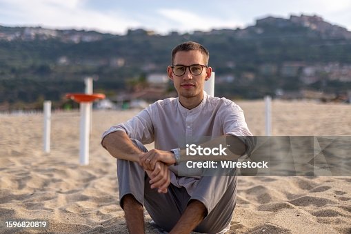 istock Young man in a relaxed pose on a sandy beach, with a tranquil ocean in the background 1596820819