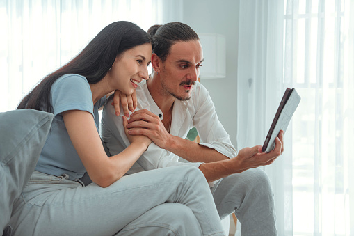 Affectionate young man embracing attractive wife girlfriend while looking at digital tablet screen, Male and female relaxing on comfortable couch. Loving couple shopping online on application