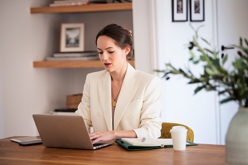 Attractive young business woman using laptop while sitting at desk and having video conference at the home office. Brunette haired female wearing white blazer.